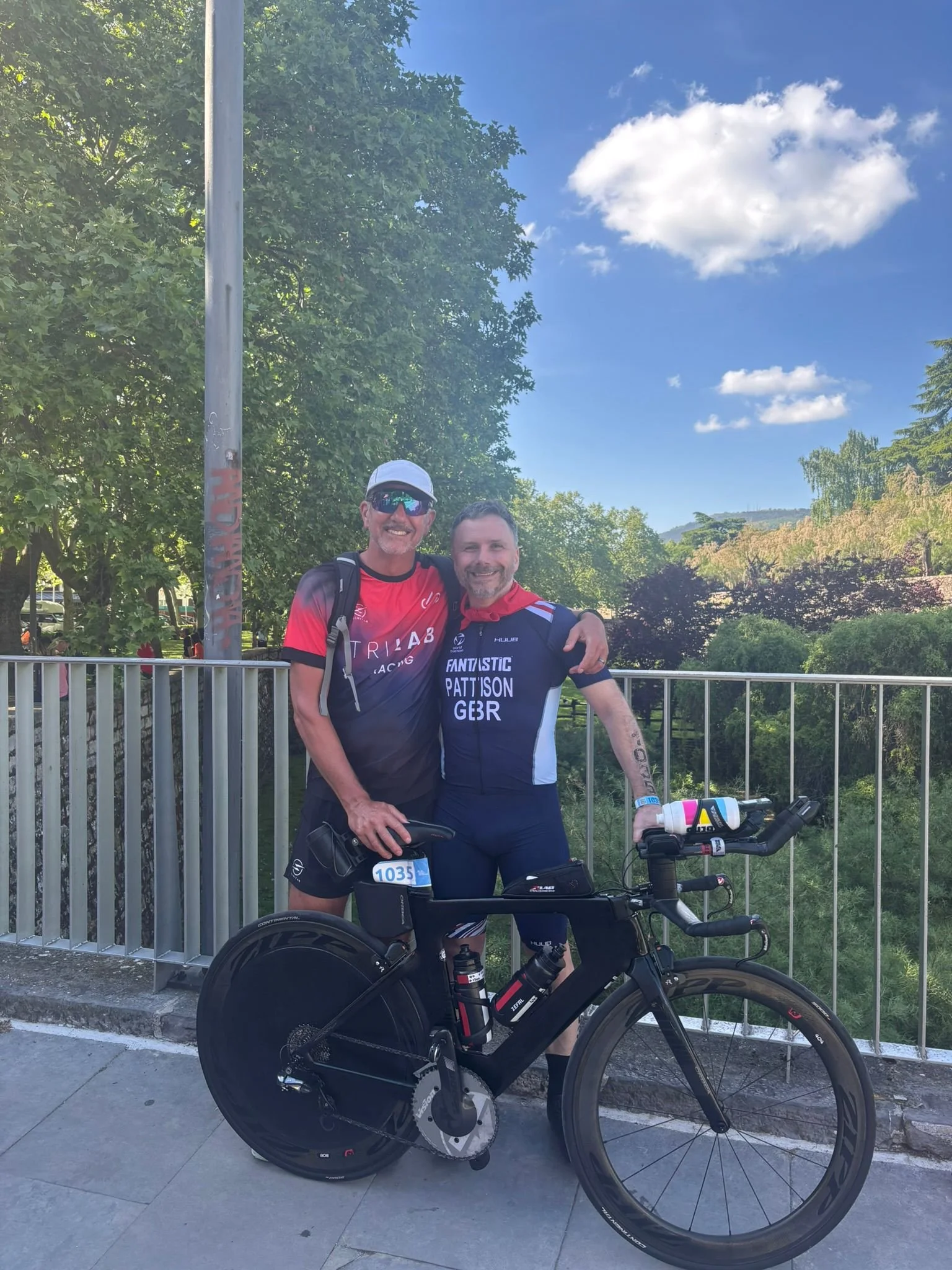 Two men in athletic gear, one with sunglasses and a cap, hugging and smiling, standing behind a black bicycle with a racing wheel, on a paved sidewalk with a metal railing, trees, and a blue sky with clouds in the background.