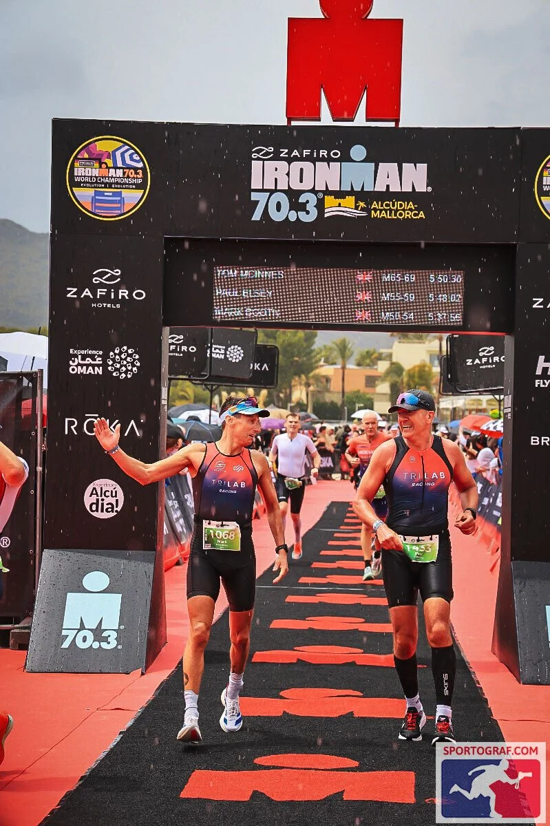 Two athletes crossing the finish line at the Ironman 70.3 in Alcudia, Mallorca, smiling and celebrating their race.