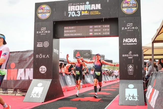 Two male athletes crossing the finish line at the Ironman 70.3 Mallorca race, with a digital timer displaying race times above the arch, spectators in the background, and race branding on the arch and surrounding posters.