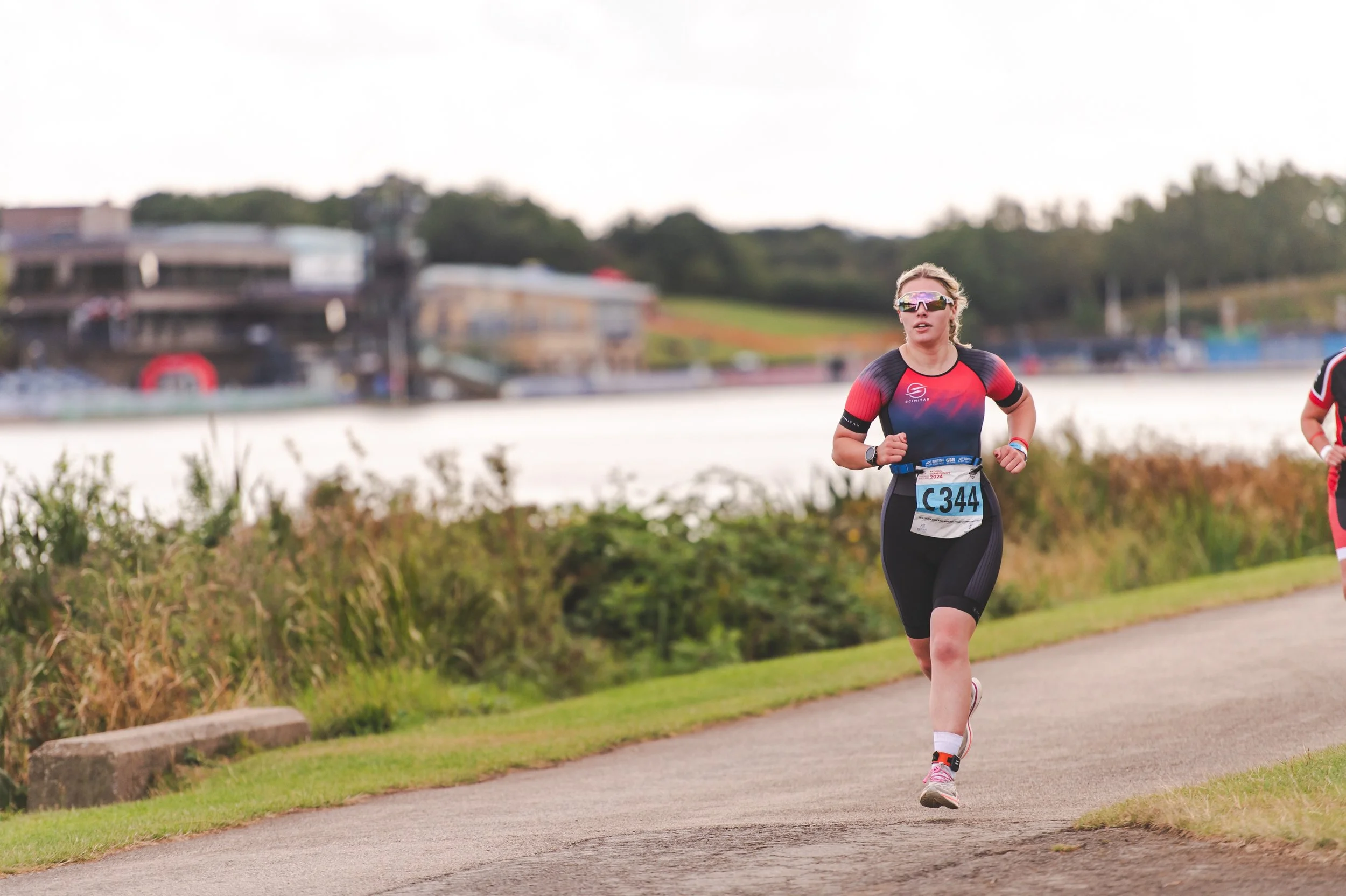 A woman running outdoors along a paved path near a body of water with trees and buildings in the background, wearing athletic clothing, sunglasses, and a race bib numbered C344.