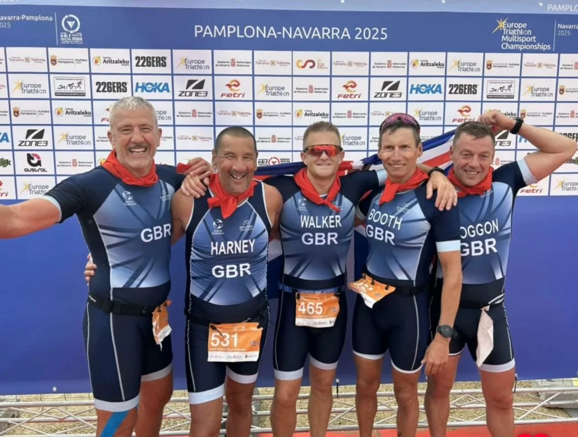 Five male athletes in matching blue and black uniforms with red neckerchiefs, standing together smiling, with arms around each other, at the Pamplona-Navarra 2025 Triathlon Championships, holding a flag, posing confidently in front of a backdrop with