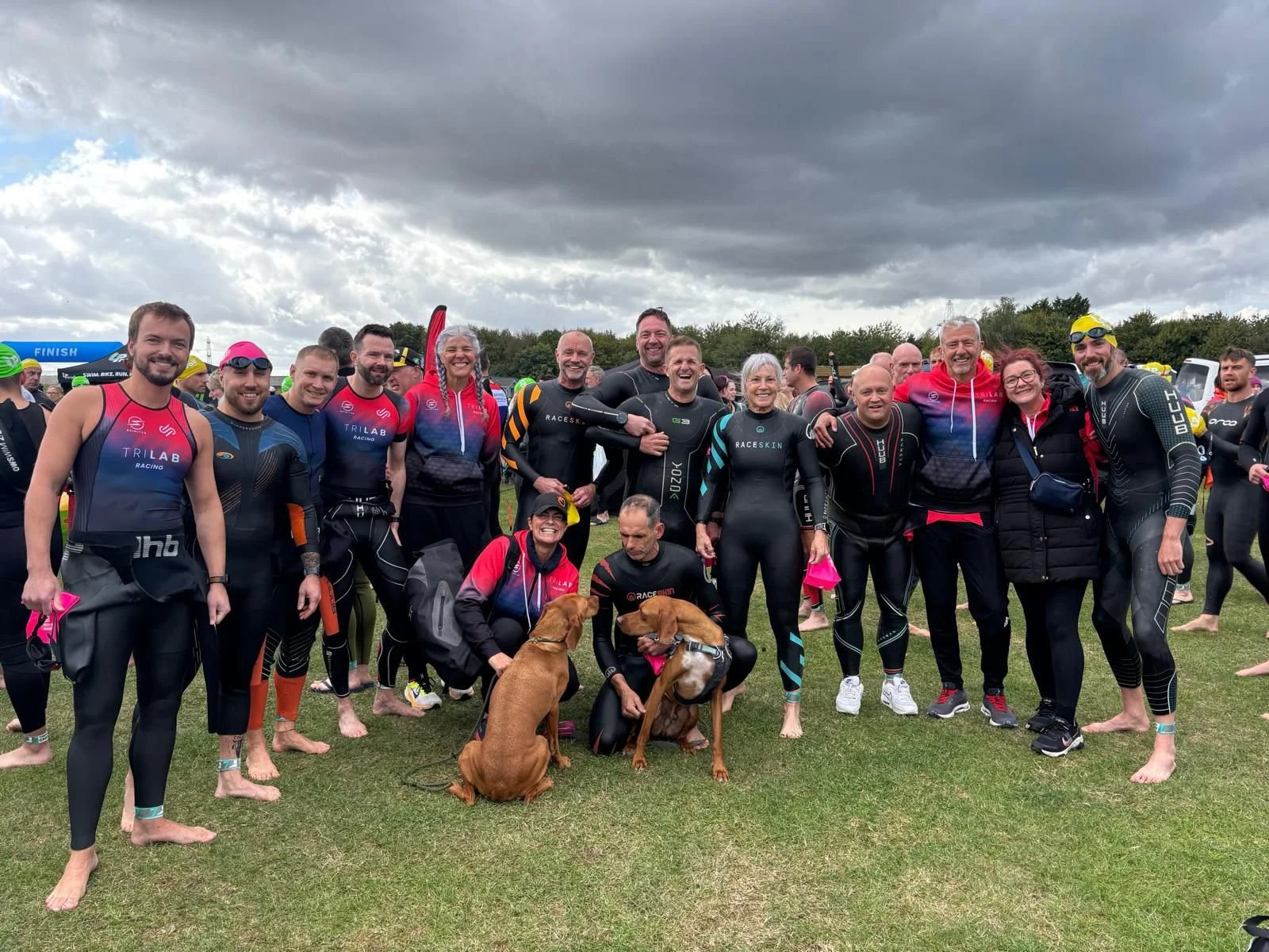Group of people in wetsuits and athletic gear at an outdoor event, some with dogs, under dark cloudy skies.