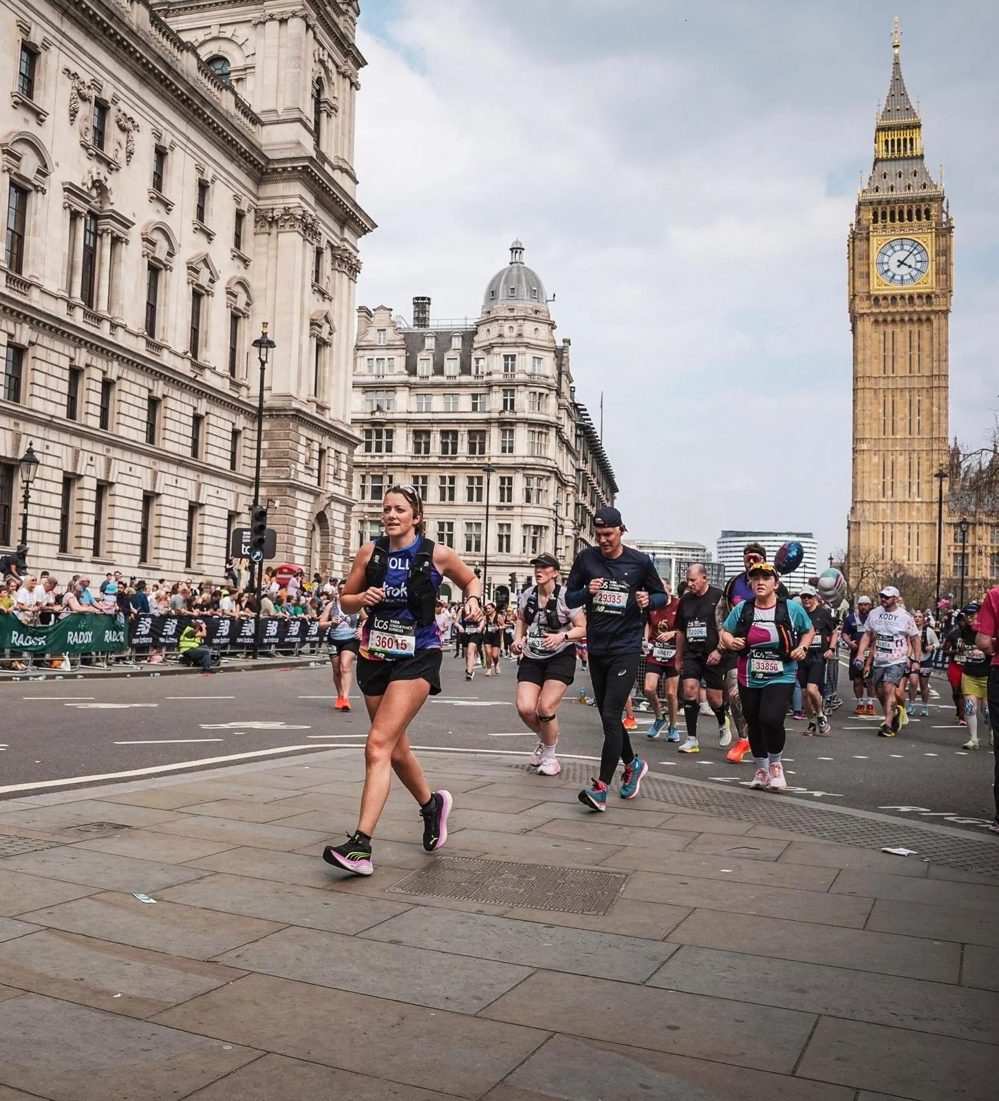 A group of marathon runners competing in a race in London with Big Ben clock tower in the background. The runners are wearing athletic clothes, bib numbers, and some have headgear or sunglasses. The street is lined with spectators and barriers.
