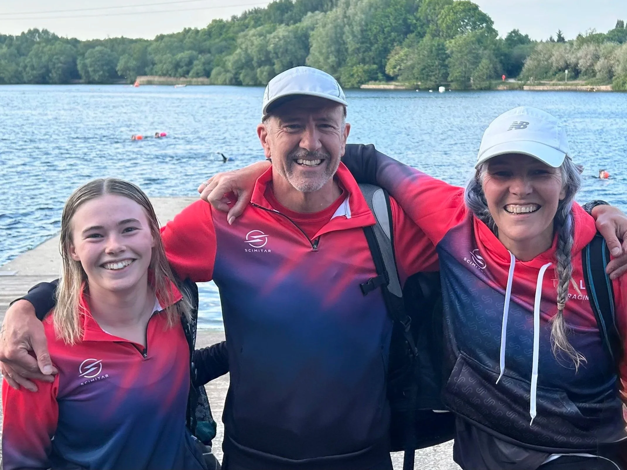 Three smiling people, two women and one man, are standing close together near a body of water with green trees in the background. They are wearing red jackets and white caps, and are outdoors on a sunny day.