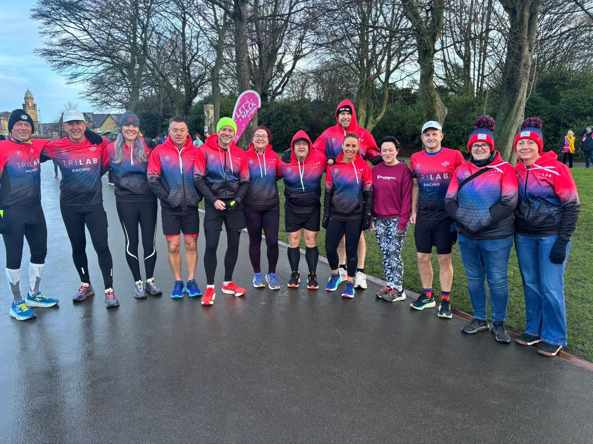 A group of runners wearing matching red and blue sports jackets standing outdoors in a park, smiling for a photo. Behind them are leafless trees and a few other people, with a cloudy sky overhead.