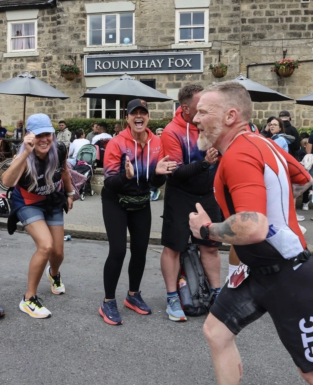 Four people in athletic clothing and running gear are outdoors, laughing and smiling, with a stone building labeled 'Roundhay Fox' in the background and others sitting at tables under umbrellas.