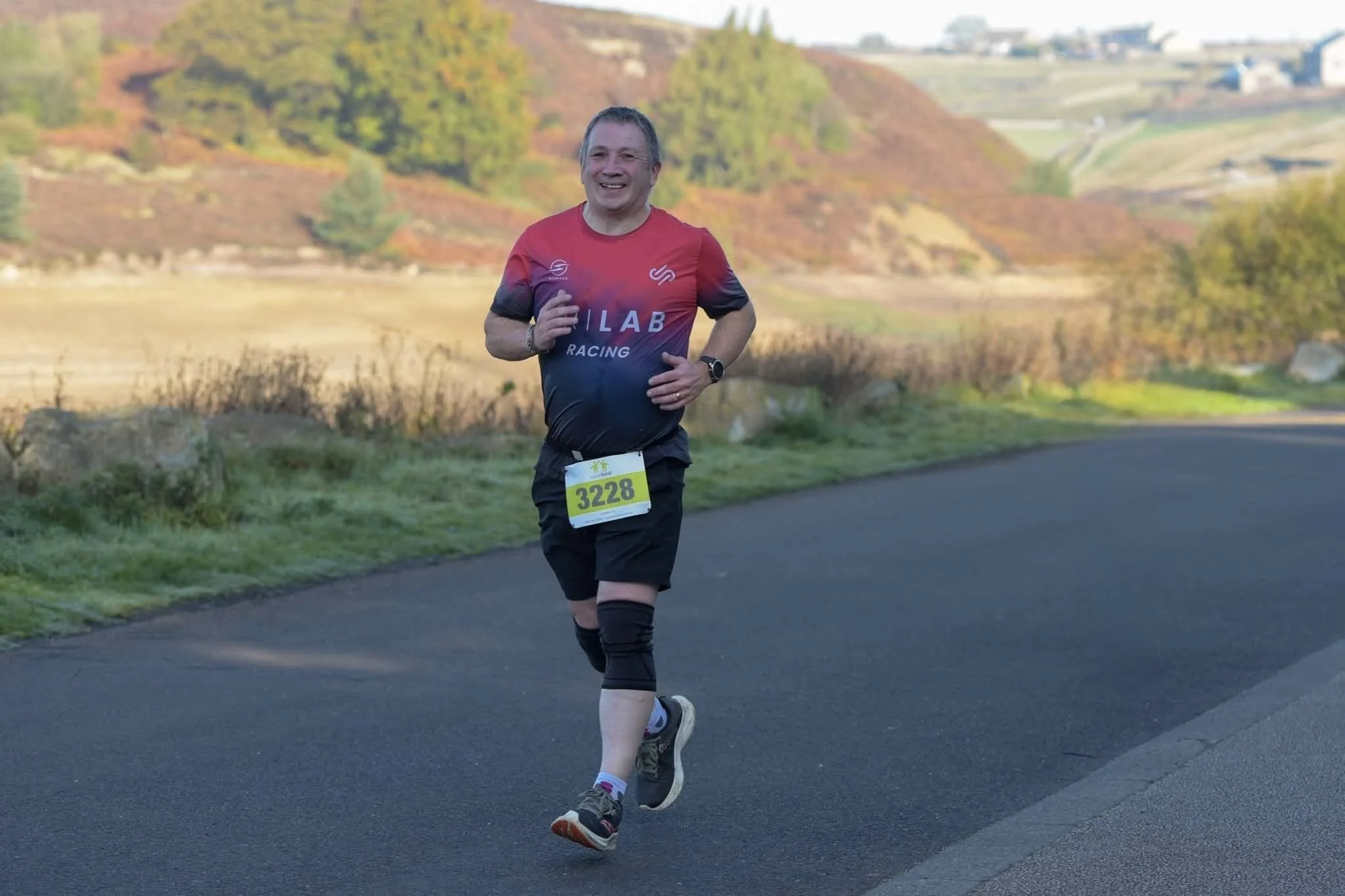 A man running outdoors during a race, wearing a red and black shirt with the words 'LAB RACING' and the number 3228 on a bib, with a scenic background of rolling hills and trees with autumn foliage.