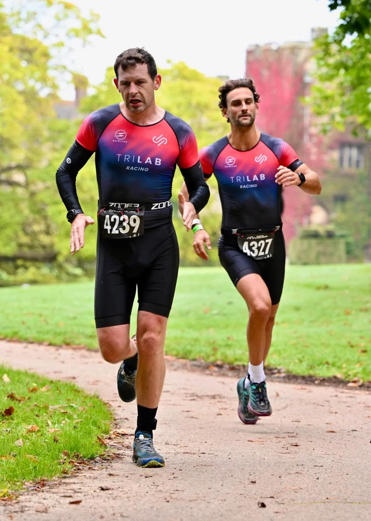 Two male runners participating in a race wearing matching black and red triathlon outfits with bib numbers 4239 and 4327, running on a park trail with green trees and grass in the background.