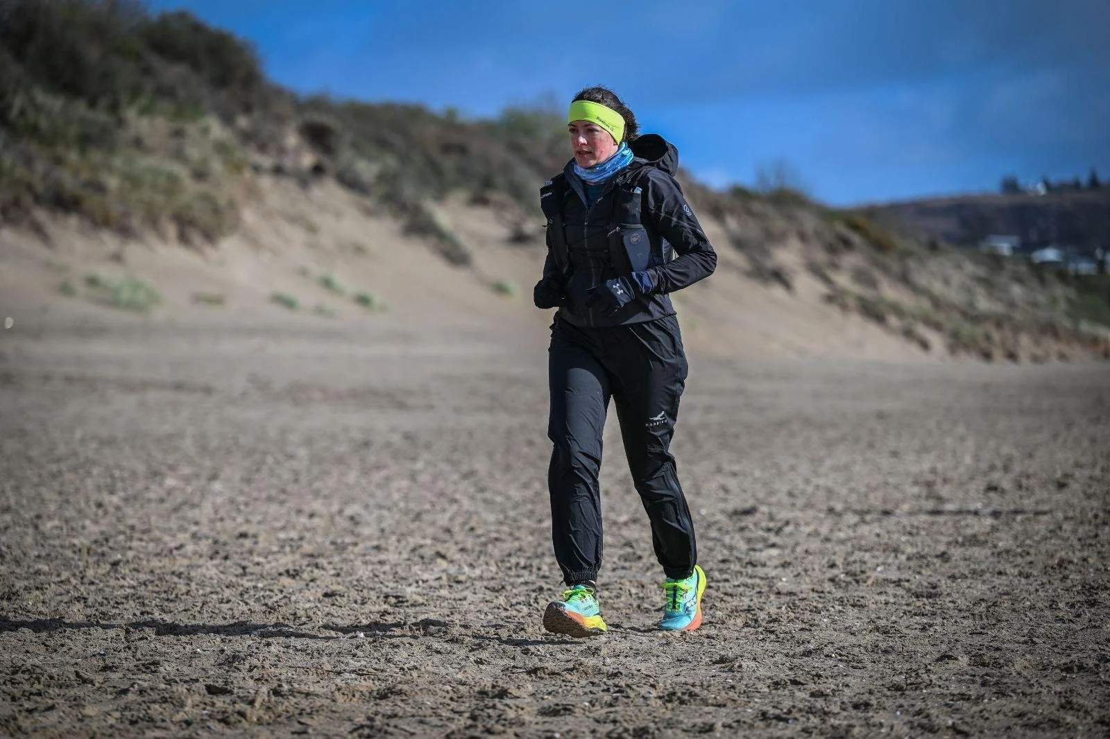 A woman running outdoors on a sandy, open area with hills in the background, wearing athletic gear including a jacket, gloves, headband, and running shoes.