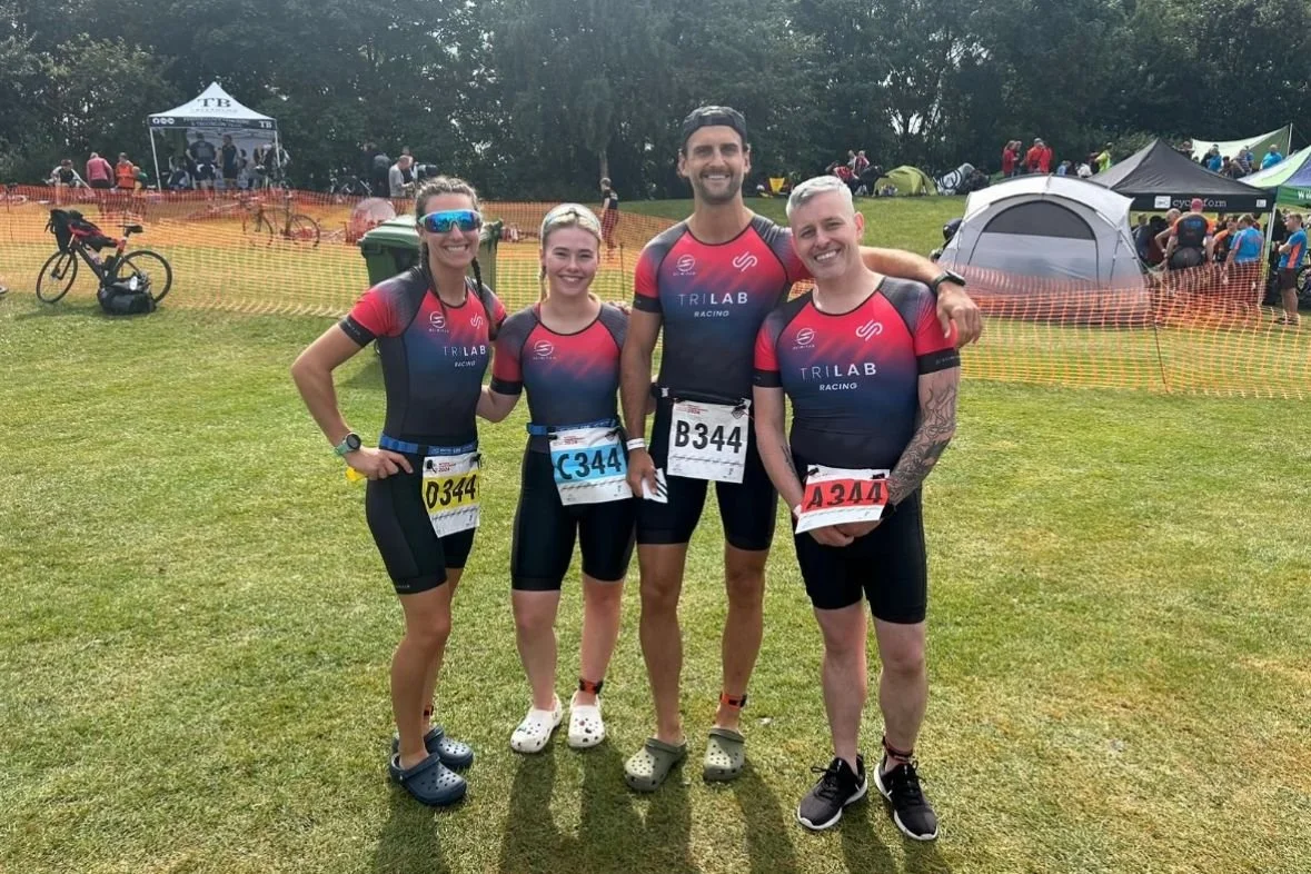 Four people in triathlon gear standing on grass at an outdoor event, smiling, with tents and groups of people in the background.