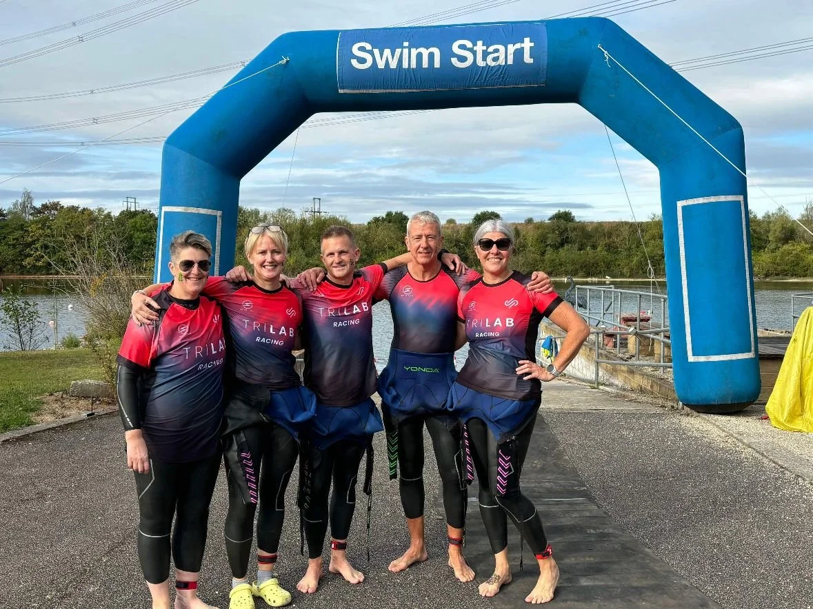 Group of five people in wetsuits standing under a blue archway labeled "Swim Start" at a lakeside open water swim event.