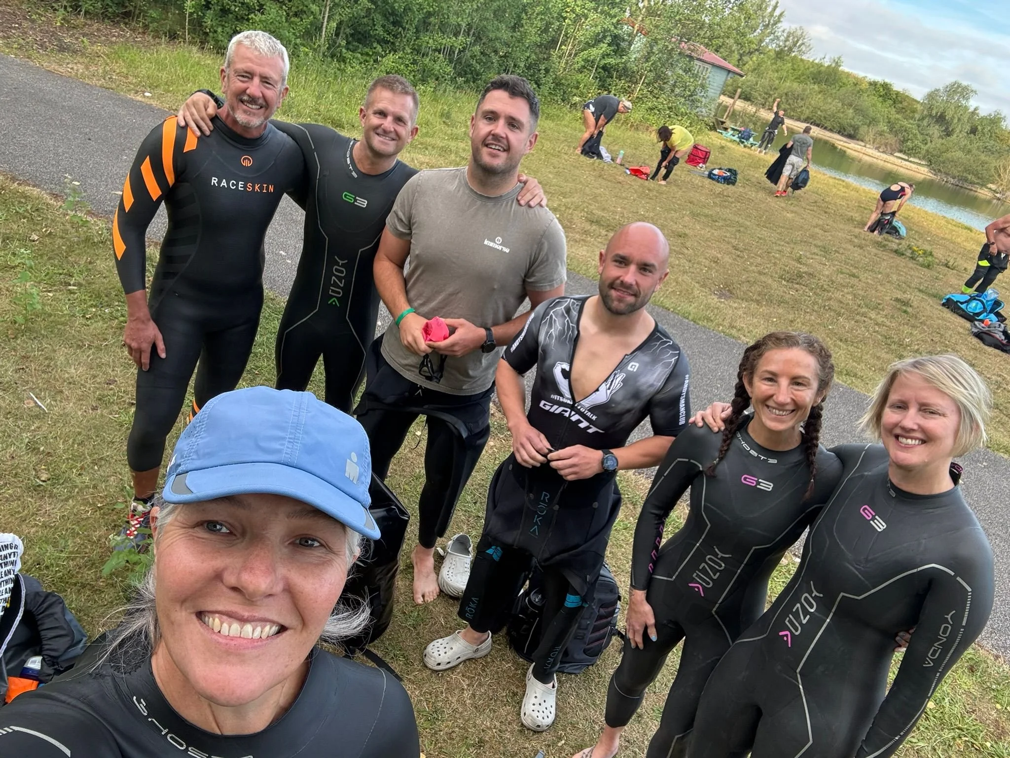Group of people standing outdoors near a lake, some in athletic wear, smiling for a selfie on a cloudy day.