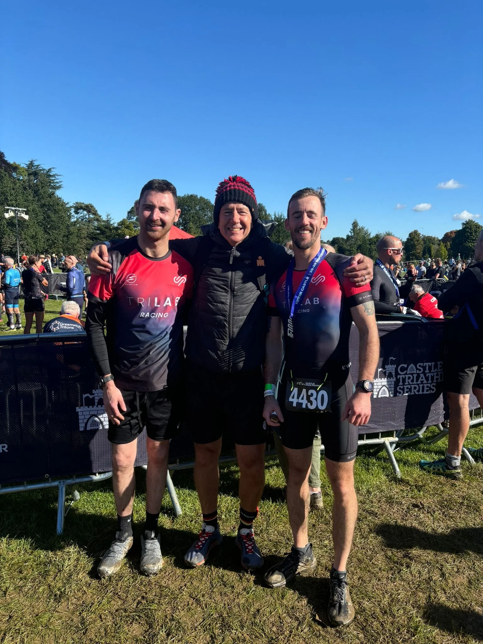 Three men standing together outdoors after a race, with a crowd and trees in the background. They are smiling, dressed in athletic gear, and one is wearing a medal.