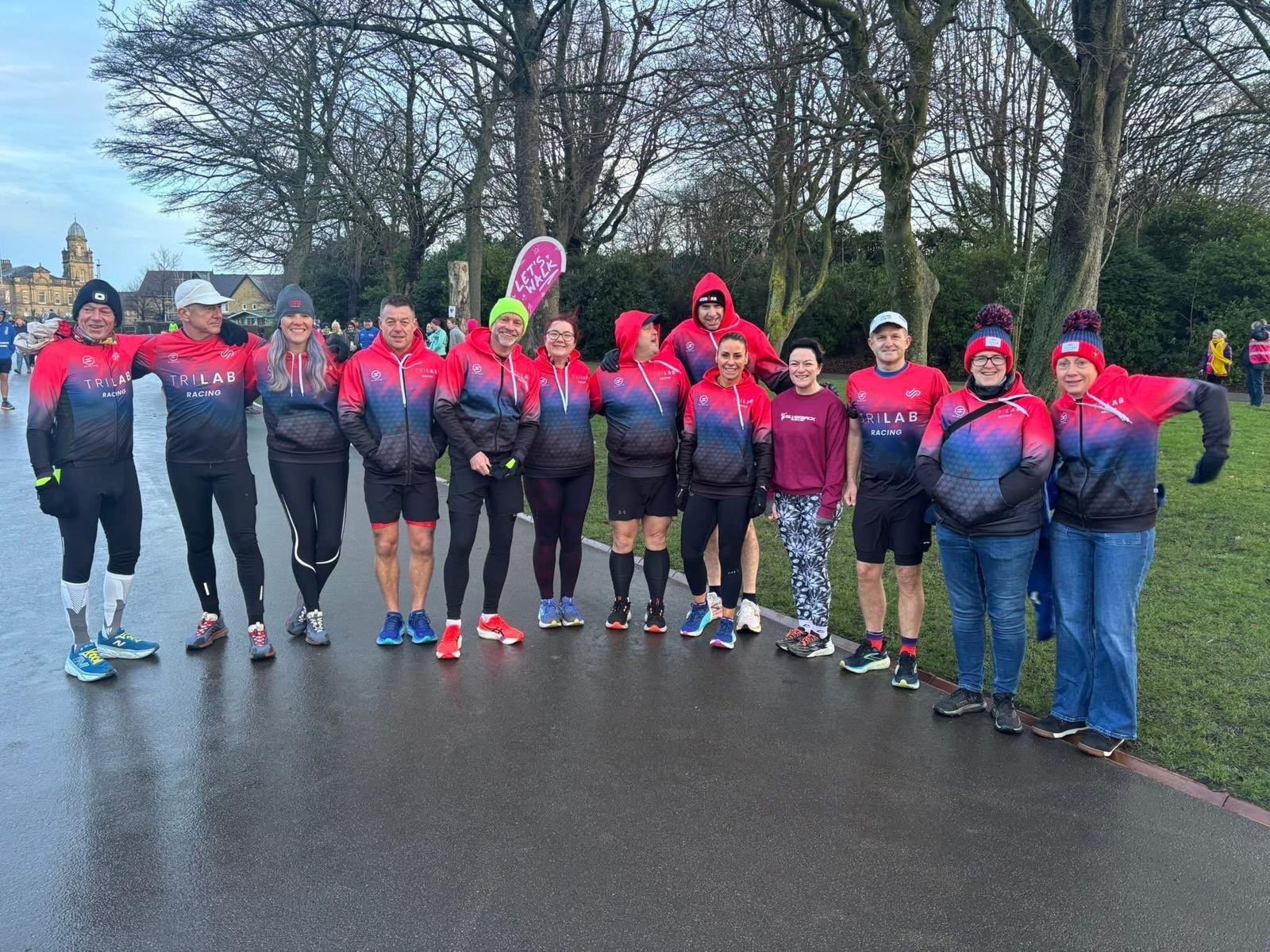 A group of people dressed in running gear standing outdoors on a cloudy day, with trees and a building in the background.