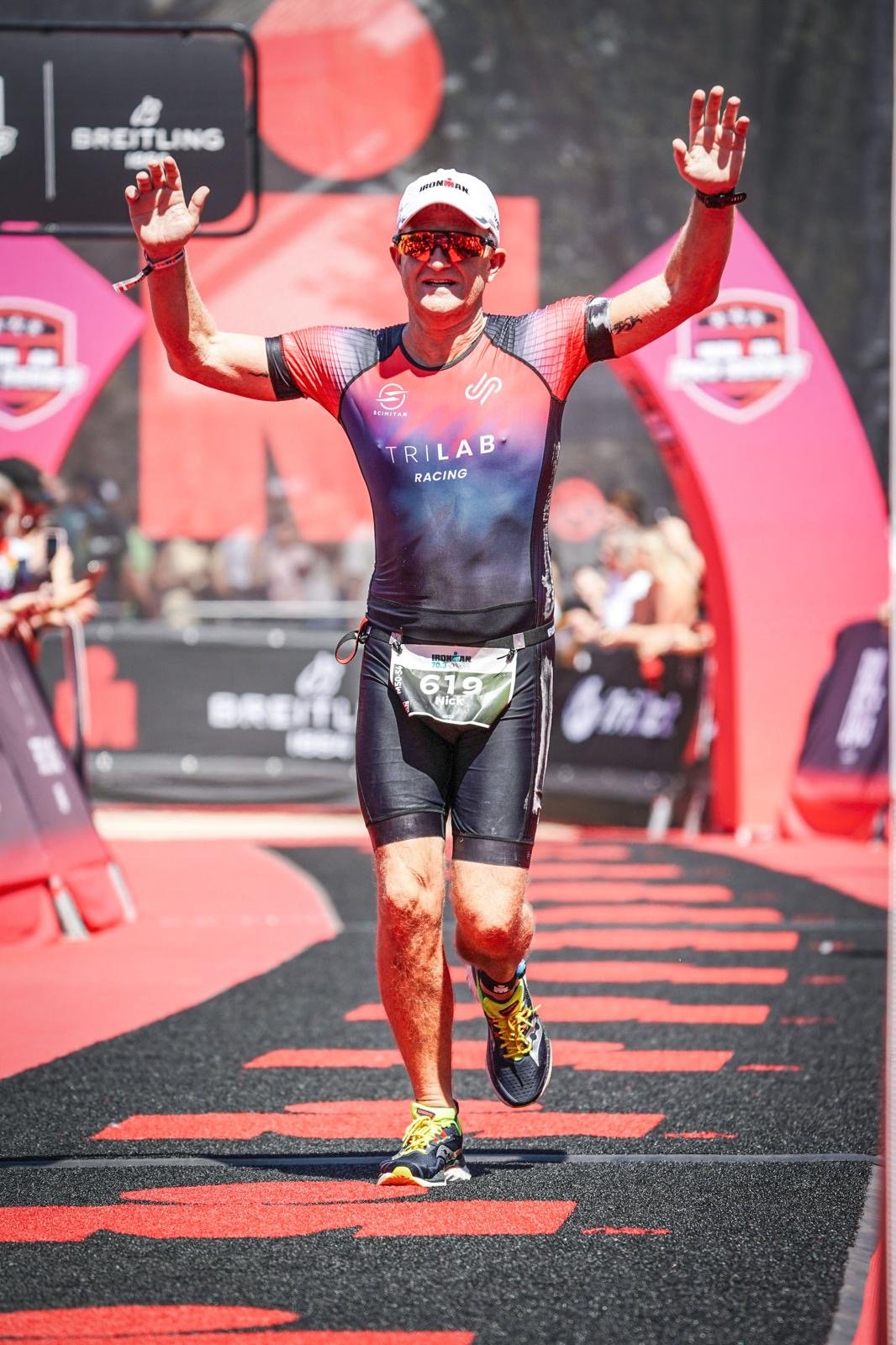 Male triathlete crossing the finish line, raising his arms in victory, wearing a black and red TriLab Racing triathlon suit, sunglasses, and a white cap, with a race bib number 619, on a red and black pathway at an outdoor sports event.