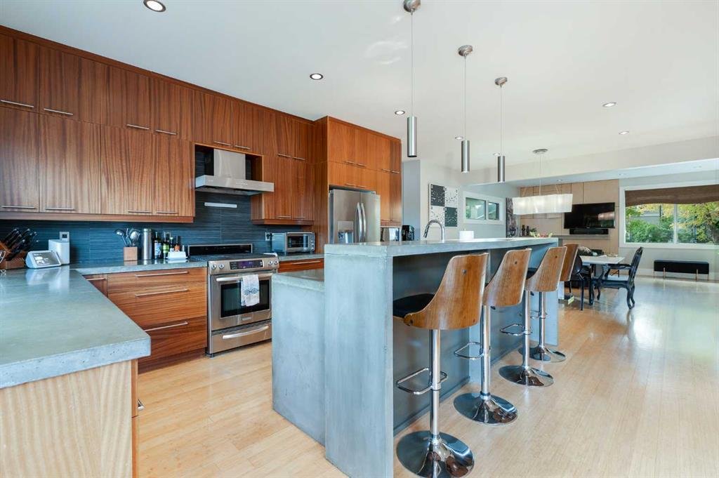 Modern open-concept kitchen with wooden cabinets, stainless steel appliances, a concrete island with four wooden barstools, and a view of the dining area and living room with large windows.