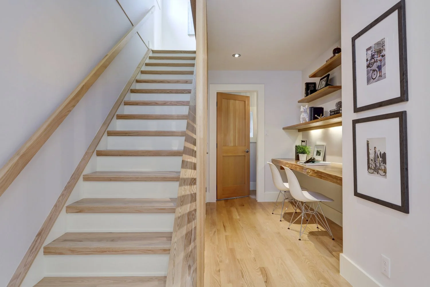 Interior view of a modern home with a staircase on the left, a small workspace with a desk and two chairs on the right, and a wooden door in the background.