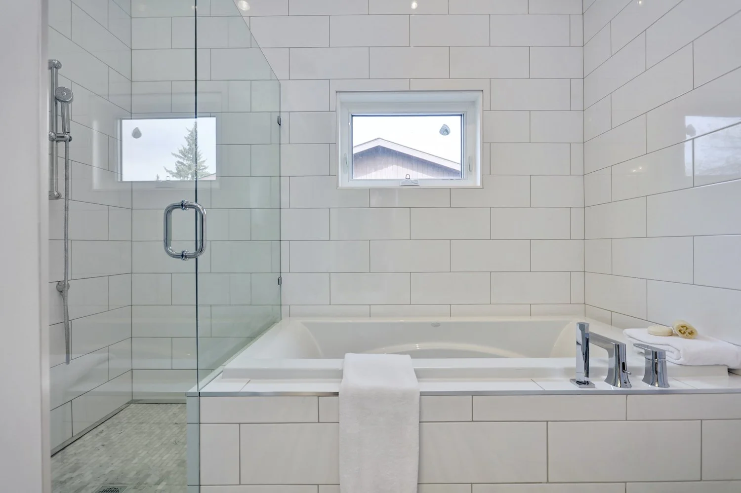 A modern bathroom featuring a white tiled bathtub with a towel draped over its edge, a small window above the tub, and a glass-enclosed shower on the left side with a showerhead and hose.