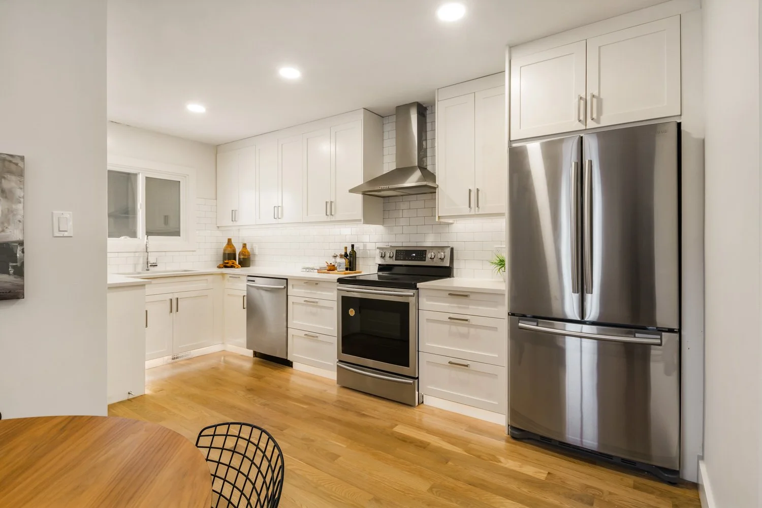Modern kitchen with white cabinets, stainless steel appliances including a refrigerator, oven, and dishwasher, white subway tile backsplash, hardwood floor, and a small wooden dining table with a black wire chair.