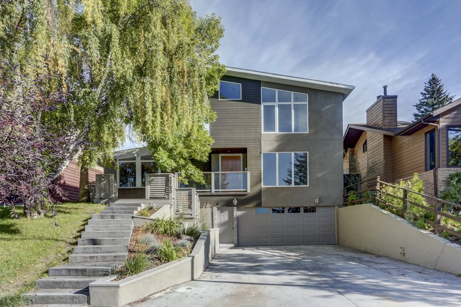 Modern three-story house with large windows, a parking garage, and landscaped stairs leading to the main entrance, with trees and neighboring houses nearby under a clear blue sky.