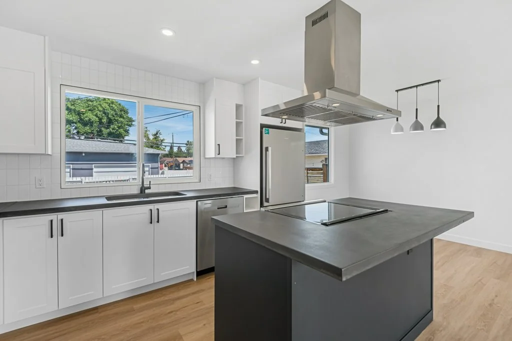 Modern kitchen with white cabinets, black countertops, stainless steel appliances, and a central island with a cooktop, pendant lights, and large windows with a view of trees and neighboring houses.