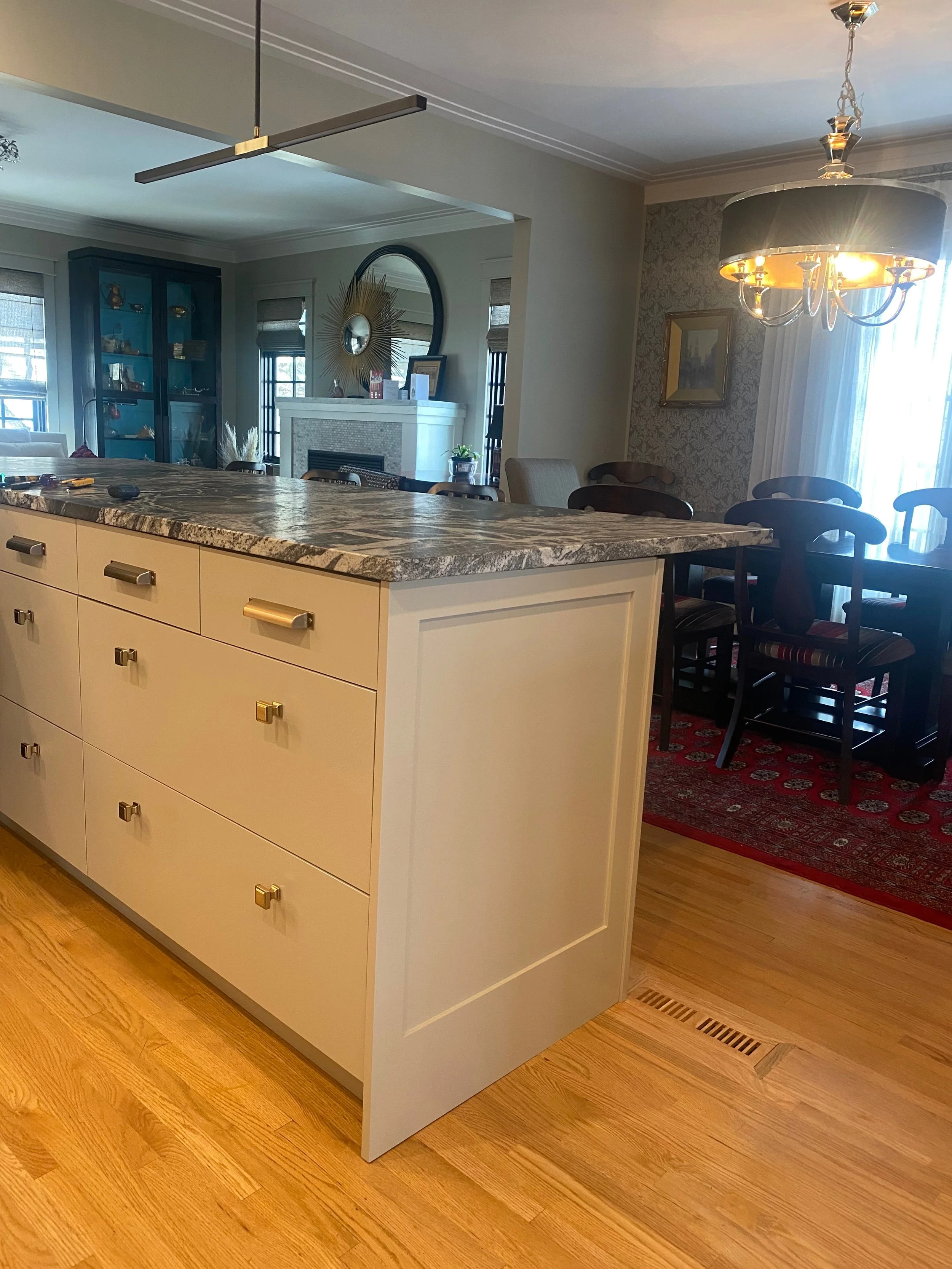 Interior view of a dining area with a kitchen island in the foreground and a dining table with chairs in the background. The island has a marble countertop and white drawers with gold and silver handles. The dining area features a chandelier, a fireplace, and a blue cabinet.