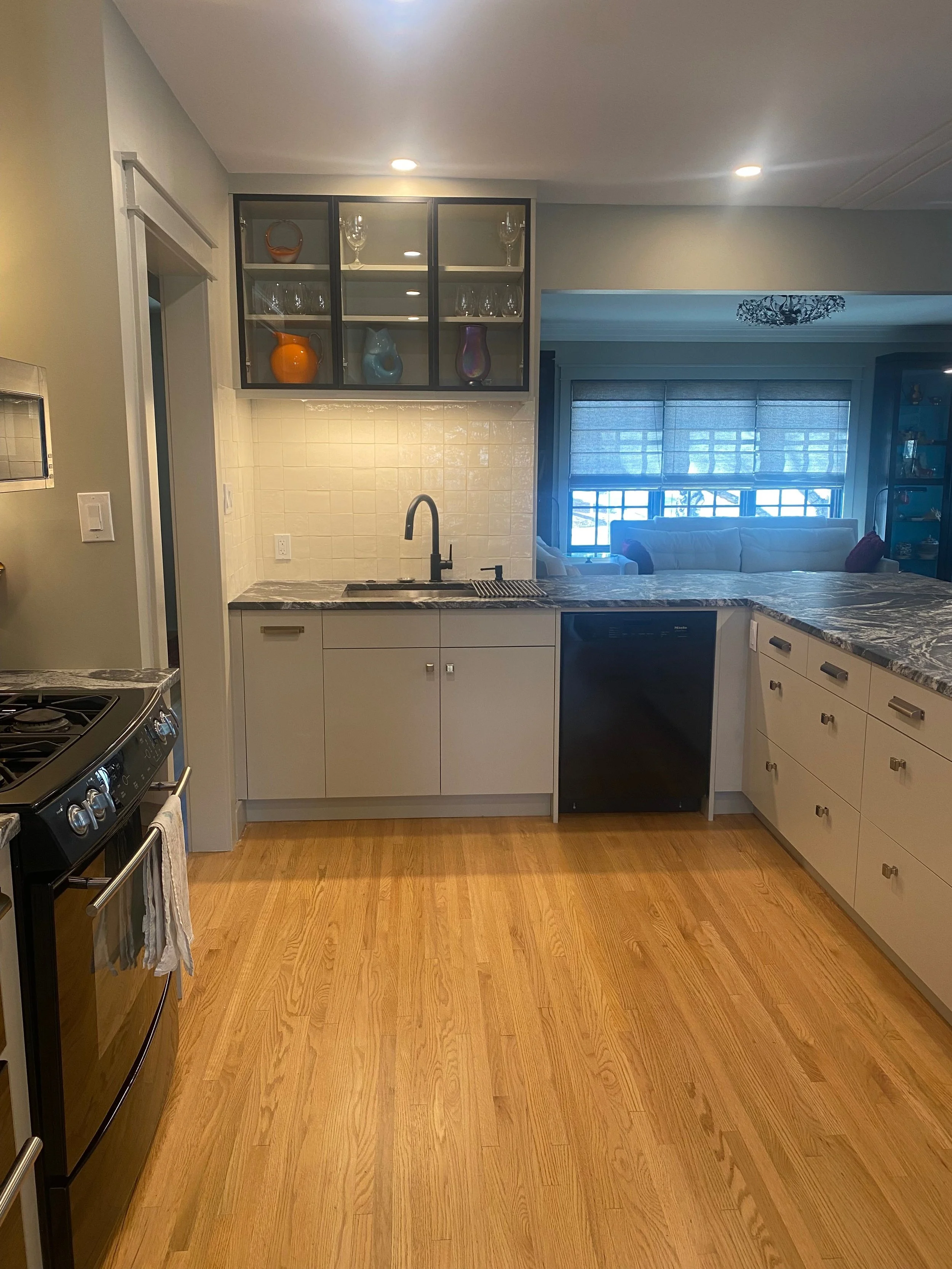 A kitchen with a black stove on the left, white cabinetry, a gray marble countertop, a sink with a black faucet, and a window with blue blinds in the background.