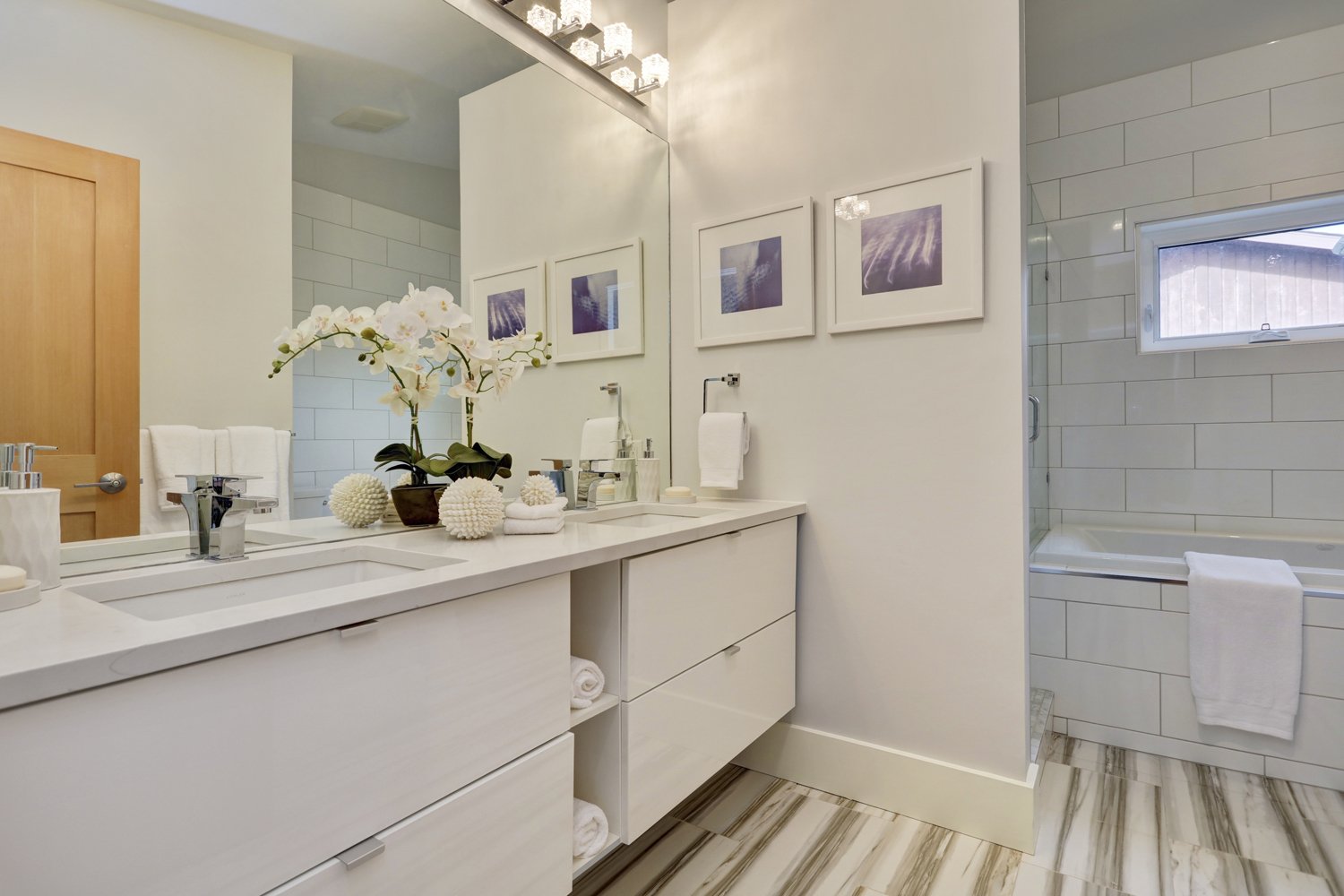 A modern bathroom with a white vanity, framed photos on the wall, a large mirror, a soap dispenser, a potted orchid, decorative balls, neatly rolled towels, a small window above the bathtub, and a towel hanging on the edge of the tub.