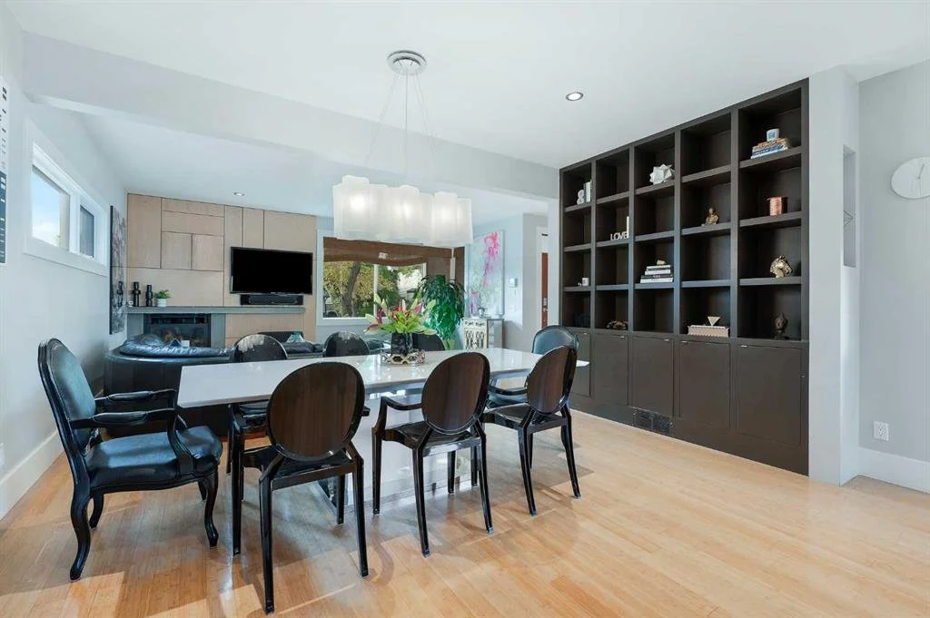 Dining room with a large black bookshelf, a white dining table with six black chairs, a chandelier, and a living area with a TV and window view.