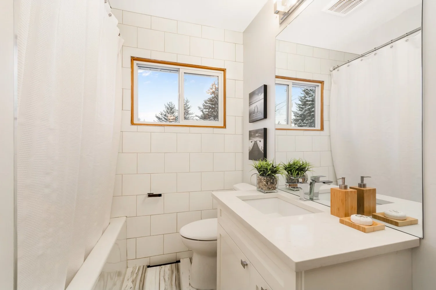 A small bathroom with white tiled walls, two small windows showing trees outside, a white bathtub with a shower curtain, a white vanity with a sink, a mirror, potted plants, and framed black-and-white pictures.