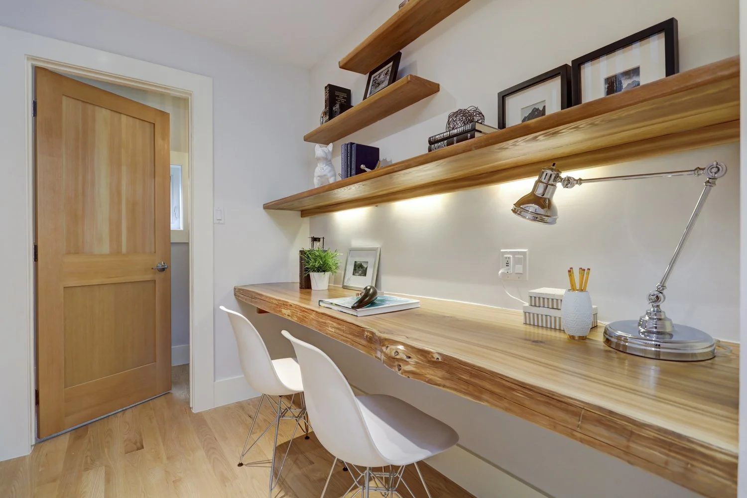 Home office with wooden desk, white chairs, open wooden shelves with books and decorations, desk lamp, and framed photos on the wall.