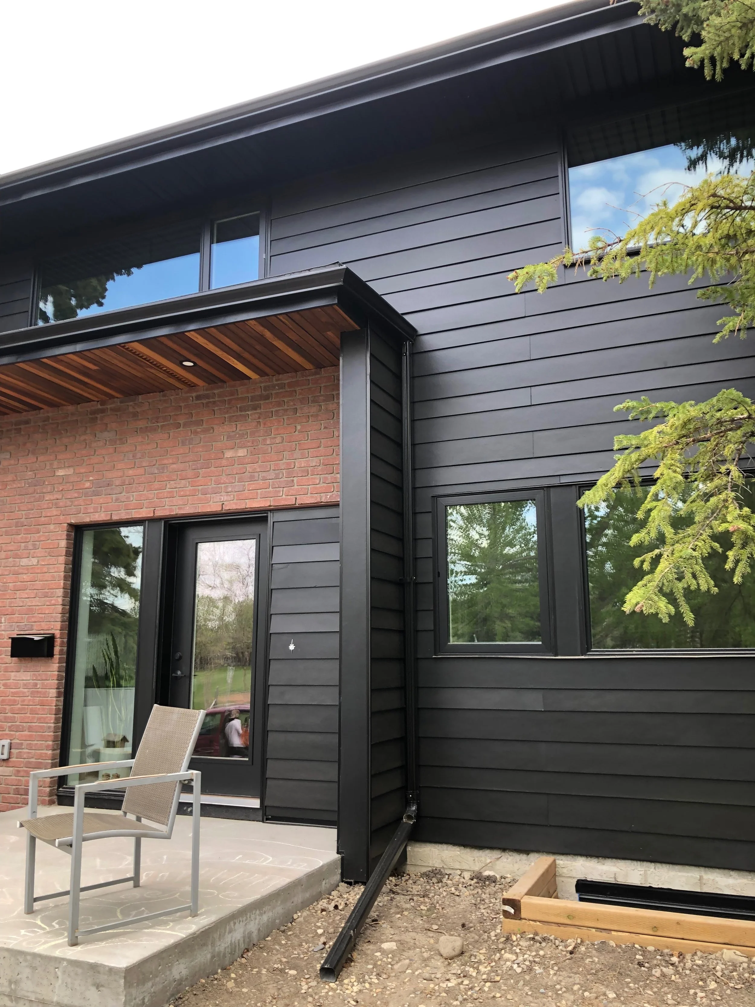 Exterior of a modern house with black siding, brick accents, a patio with a chair, and windows reflecting trees.