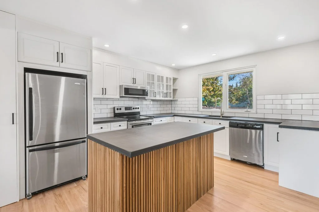 Modern kitchen with white cabinets, stainless steel appliances, black countertops, a central island with wood paneling, and a window overlooking trees.