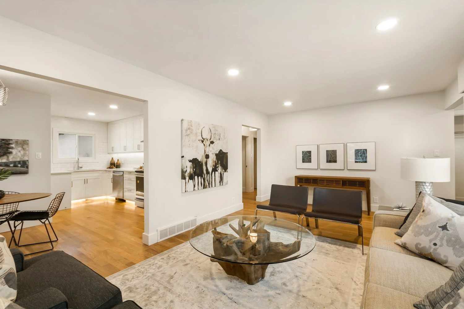 Living room with a beige sofa, black chairs, a glass coffee table with a wooden base, art on the white walls, wooden flooring, and a view into the kitchen.