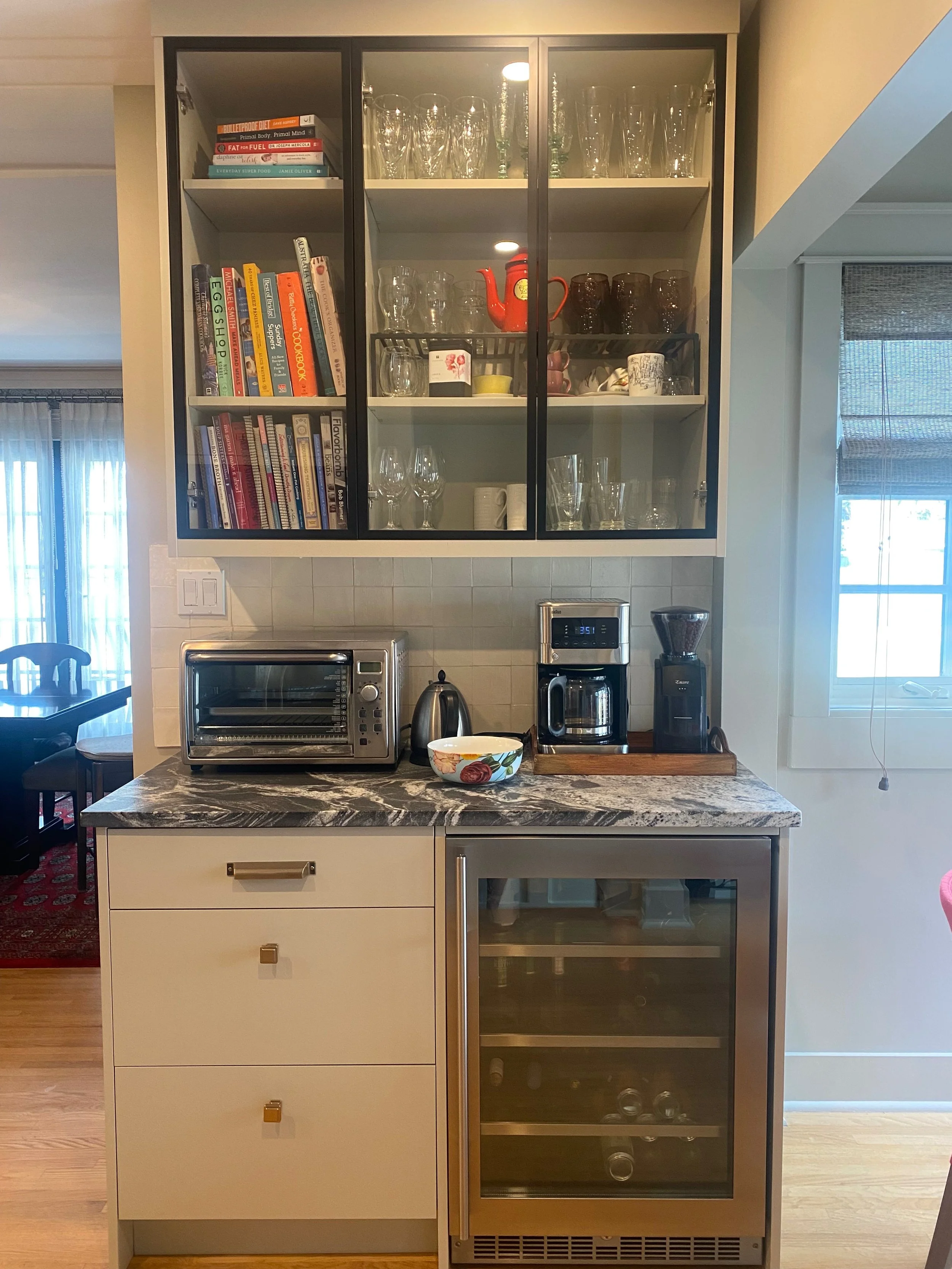 Kitchen counter with toaster oven, electric kettle, coffee Maker, and wine fridge. Cabinet above with glasses, cups, books, and decorative items.