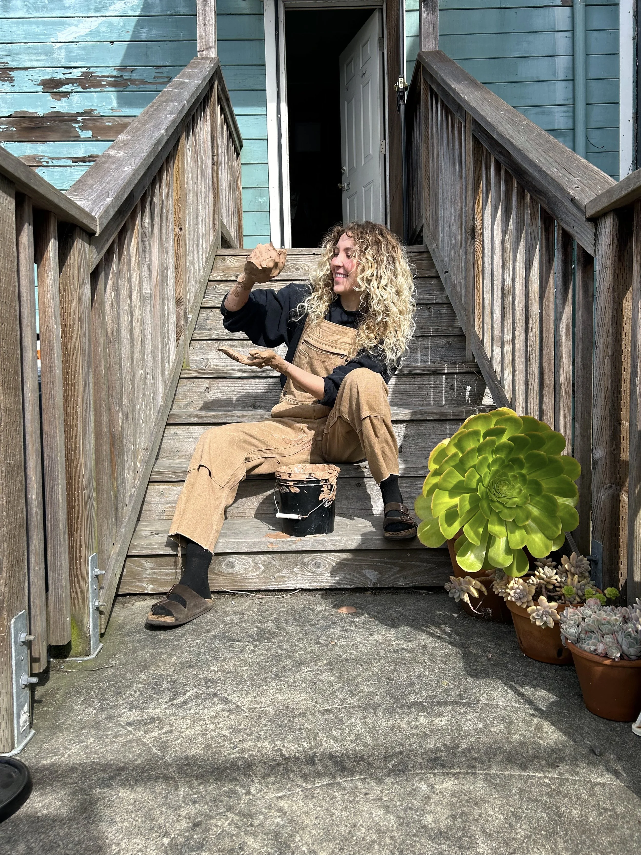 A young woman with a halo of curly blonde hair sits on the wooden back steps of a well-worn shop painted light aqua. She's wearing a black sweatshirt, camel overalls, & is playing with a mess of wet clay. Next to her is a big succulent plant!