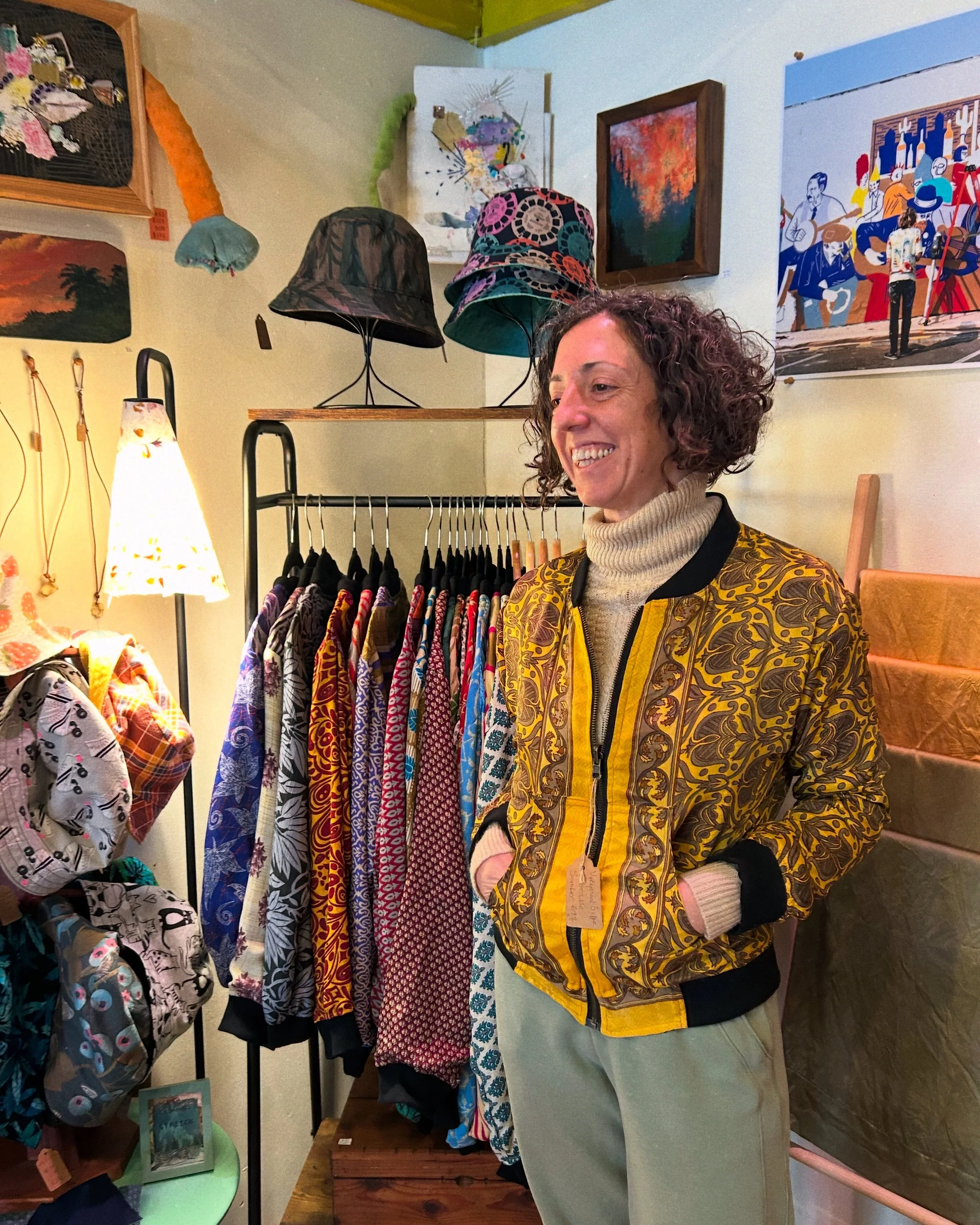 Maria, an ethnically ambiguous middle-aged woman with dark curly hair cut into a bob, stands smiling in front of a clothing rack displaying dozens of the upcycled sari bomber jackets she makes. She is modeling a bold yellow and black jacket.
