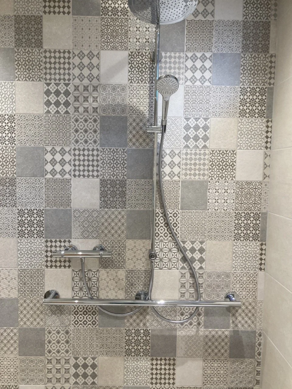 Ein Duschkopf an einer Wand mit Musterfliesen in Grau- und Beige-Tönen.