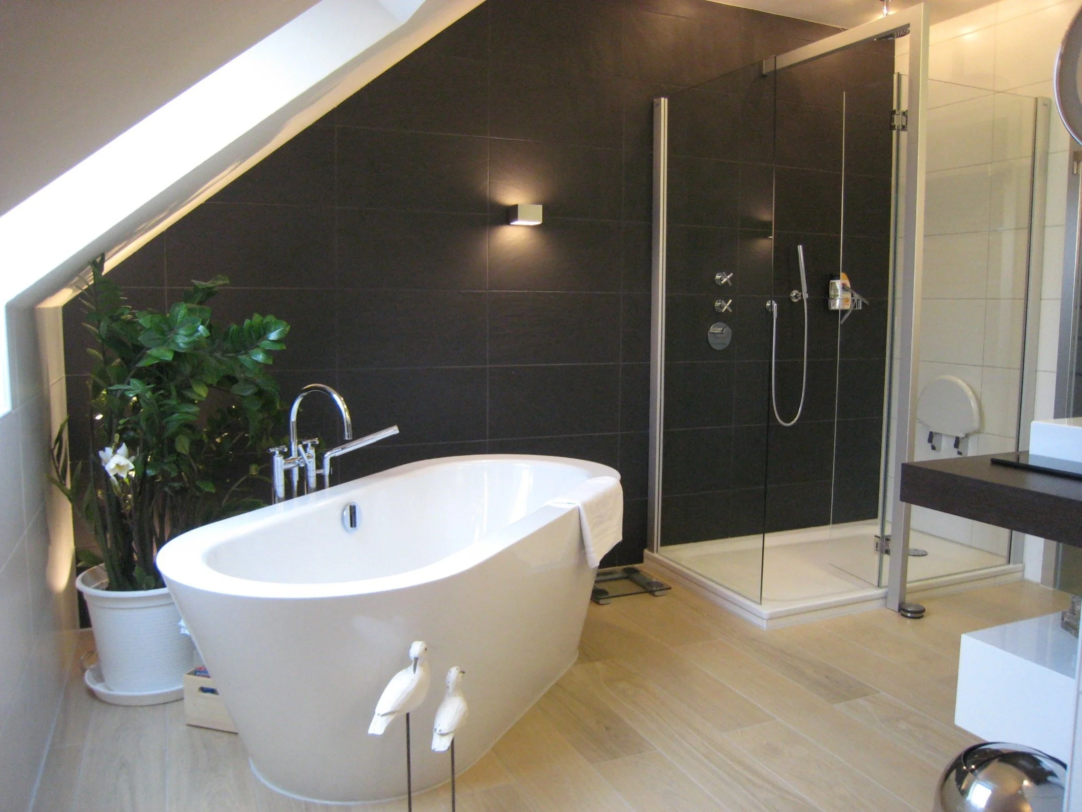 Ein modernes Badezimmer mit freistehender Badewanne, einer Duschkabine, einer Pflanze in einem weißen Topf, Holzfußboden und dunklen Wandfliesen.