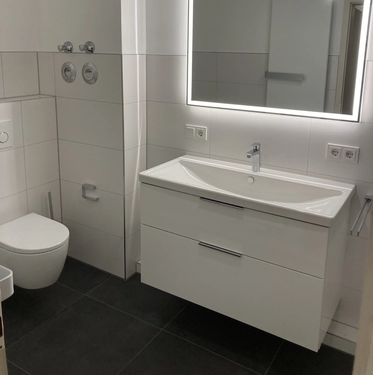 Modernes Badezimmer mit weißen Fließen, einer Spiegelbeleuchtung, Waschbecken mit Schrank, einem WC und mehreren Steckdosen.