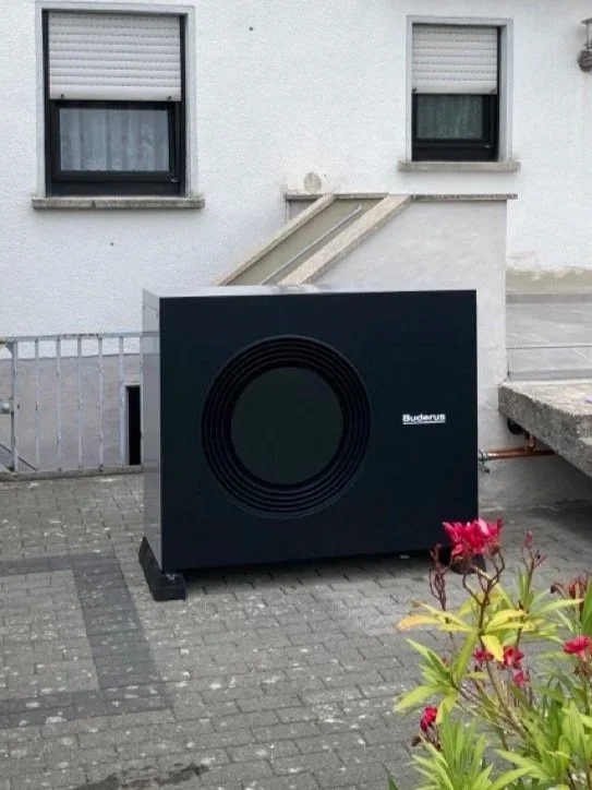 Außenansicht eines Gebäudes mit zwei Fenster und einer großen schwarzen Heizungs- oder Heizkesselanlage mit dem Label 'Buderus' vor einem gepflasterten Bereich und Blumen im Vordergrund.