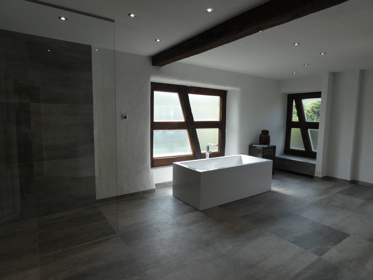 Ein modernes Badezimmer mit freistehender Badewanne, dunklen Holzfenstern und grauen Fliesen, hellem weißen Wänden und einer dunklen Holzdecke.