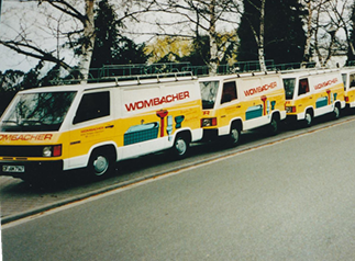 Mehrere weiße Busse mit gelber und roter Werbung für Wombacher stehen am Straßenrand.