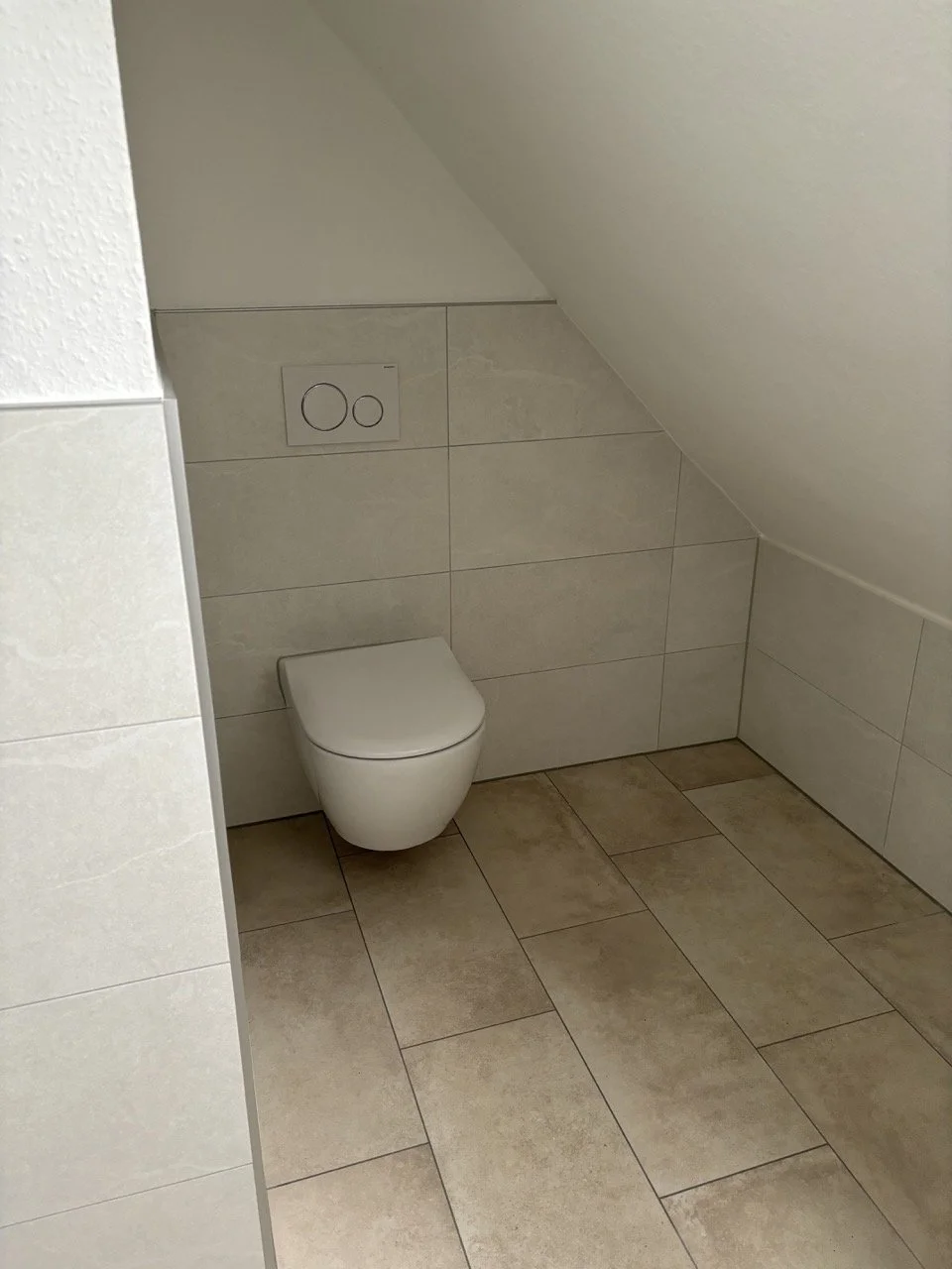 Ein Badezimmer mit einer wandhängenden Toilette und einer verdeckten Spülung, Fliesen an Wand und Boden, und einer schrägen Decke.