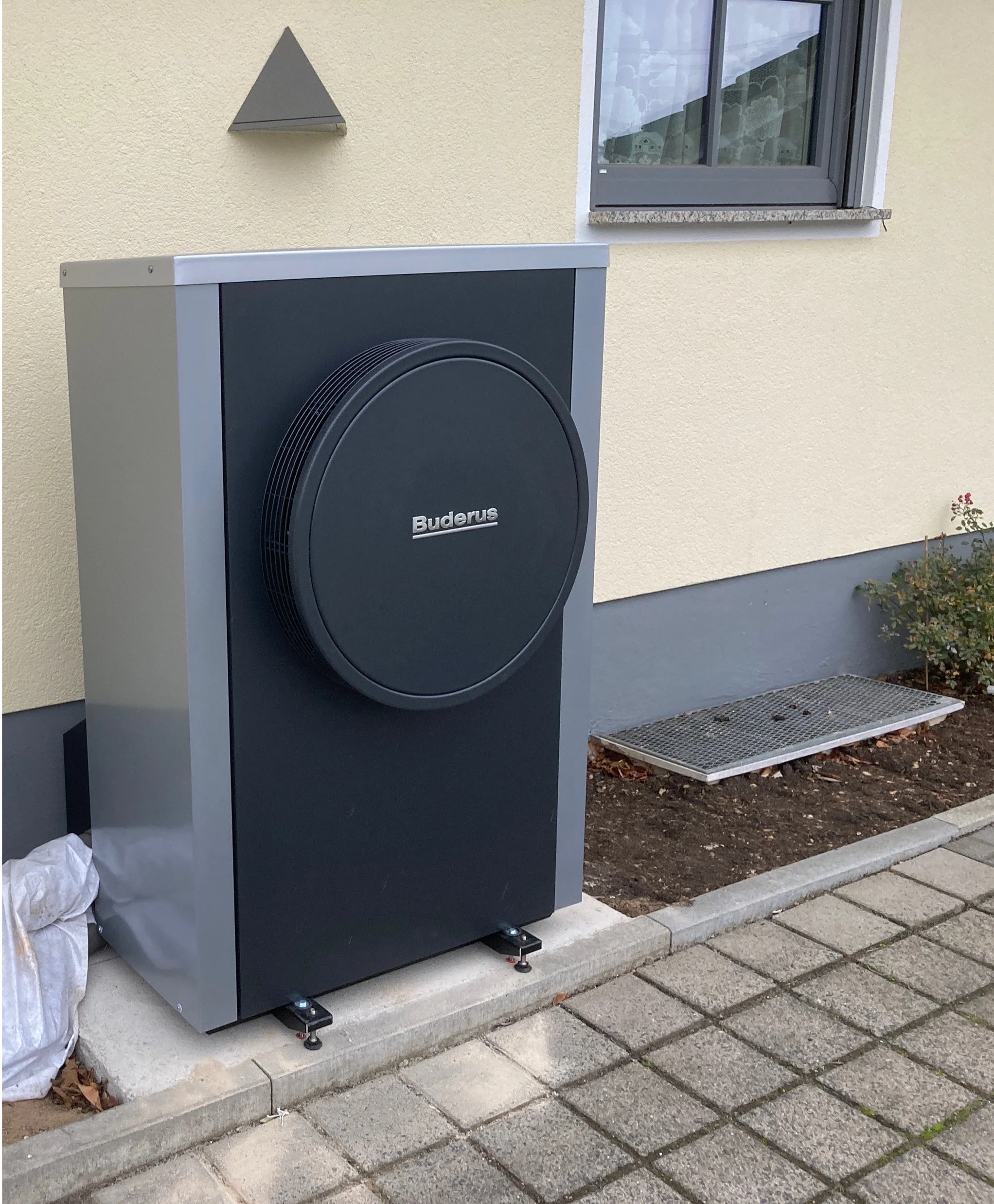 Außenansicht einer modernen Heizungsanlage mit einem großen, schwarzen Buderus-Logo vor einem Haus mit gelb- und graufarbener Fassade.