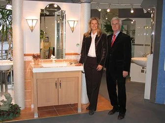 Zwei Menschen, ein Mann und eine Frau, stehen vor einem Badezimmer mit einem Waschbecken und Spiegel. Es scheint ein Ausstellungsraum für Badezimmermöbel zu sein.