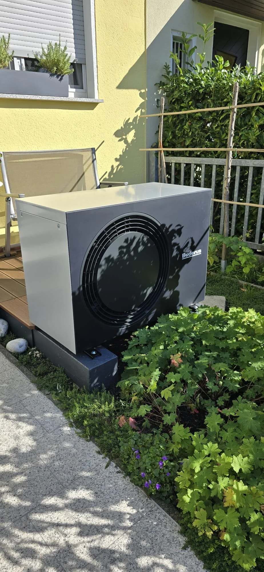 Außengerät einer Klimaanlage oder Wärmepumpe auf einer Terrasse neben Pflanzen und einem Gartenzaun, vor einem gelben Haus mit einem Fenster mit Blumenkasten.