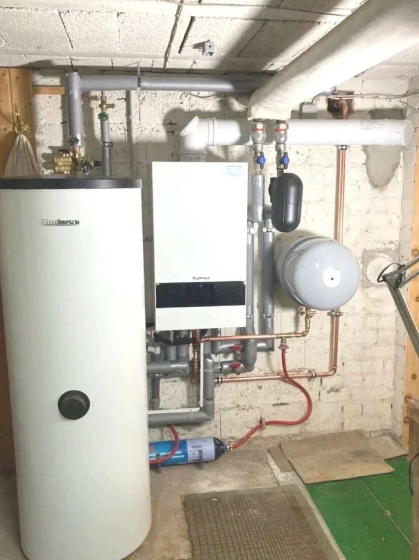 Heizungs- und Wasserboiler in einem Kellerraum mit Rohren, Pumpen und einem Ausdehnungsgefäß.