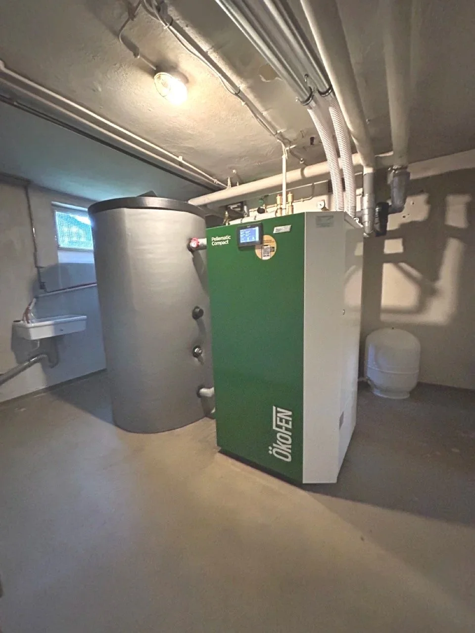 Ein grün-weißes Heizgerät namens ÖkoFEN und ein großer zylindrischer Wasserspeicher in einem Kellerraum.