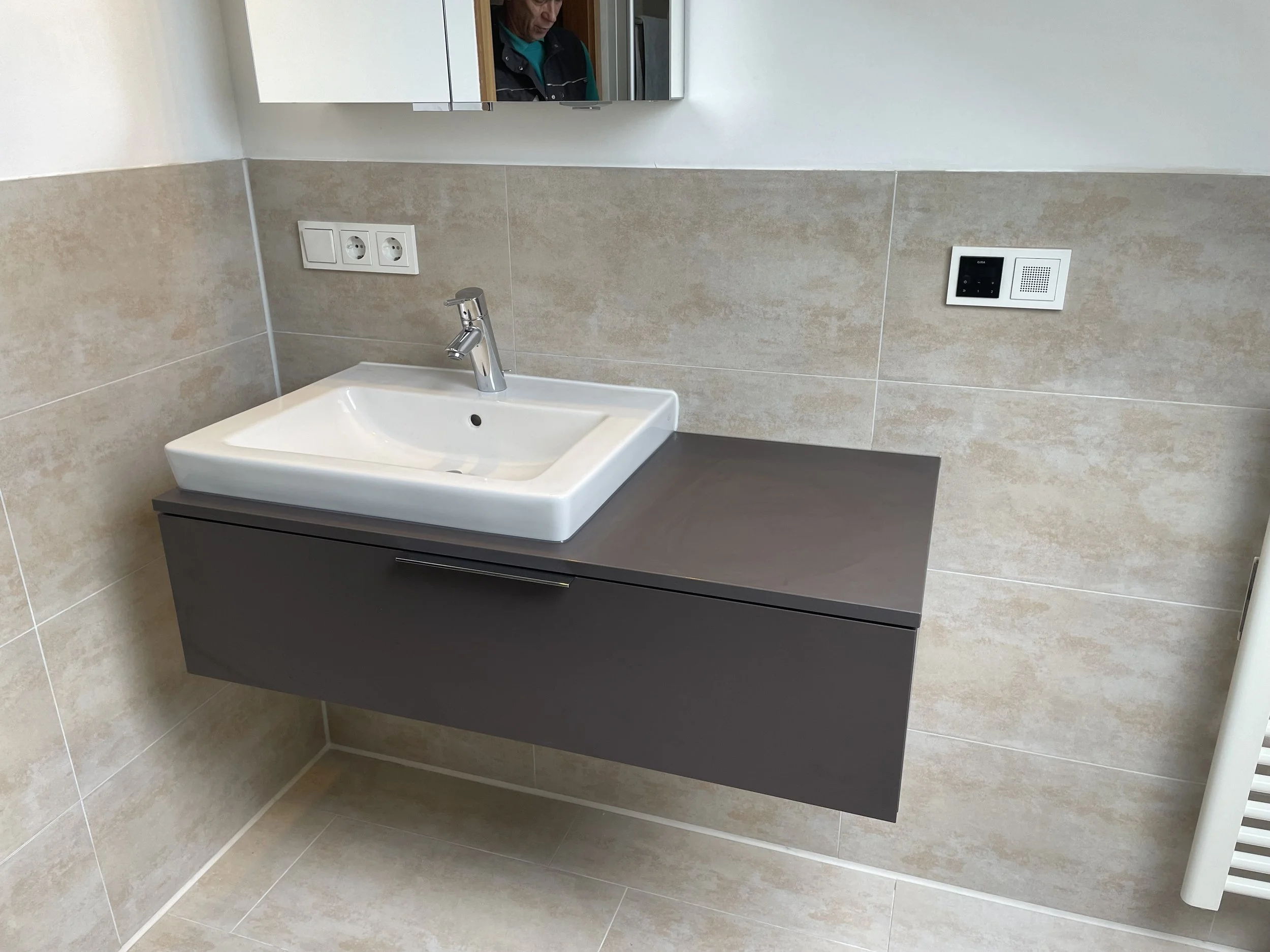 Waschbecken mit spiegel, dunkle Wandablage, elektrische Anschlüsse und Steckdosen auf einer beige gefliesten Wand.